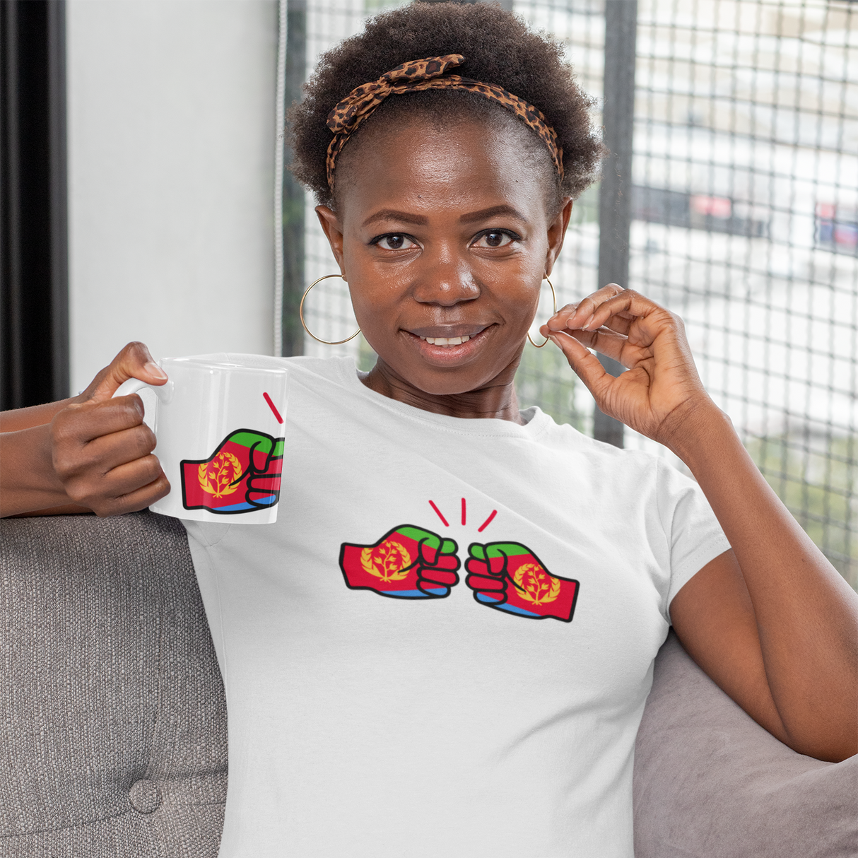 Woman wearing a white We Run Tings Eritrea Mug and t-shirt with colorful graphics, sitting on a couch.