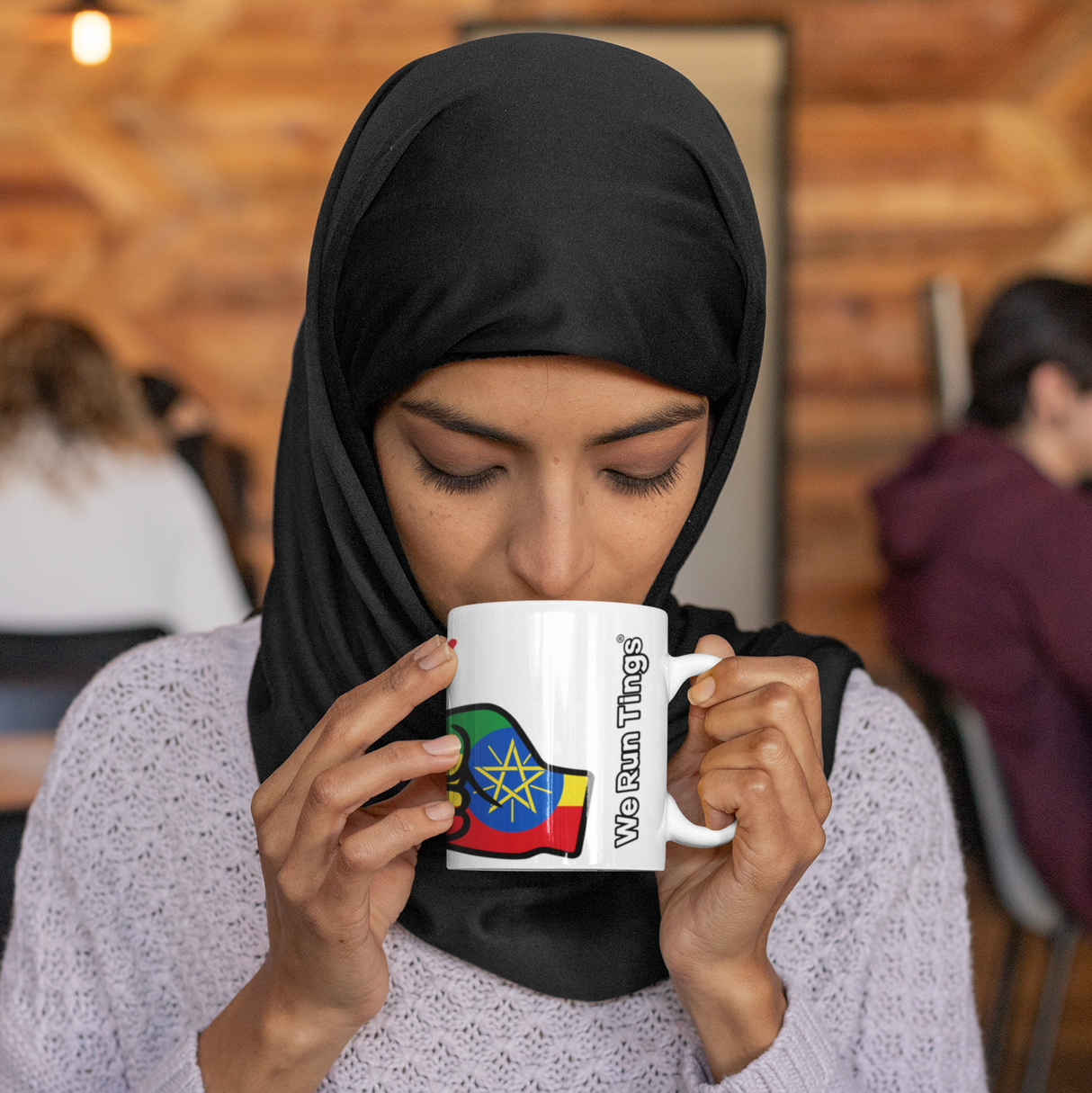 Woman wearing a hijab drinking from a We Run Tings white ceramic Ethiopia mug with a colorful design, sitting in a casual setting.