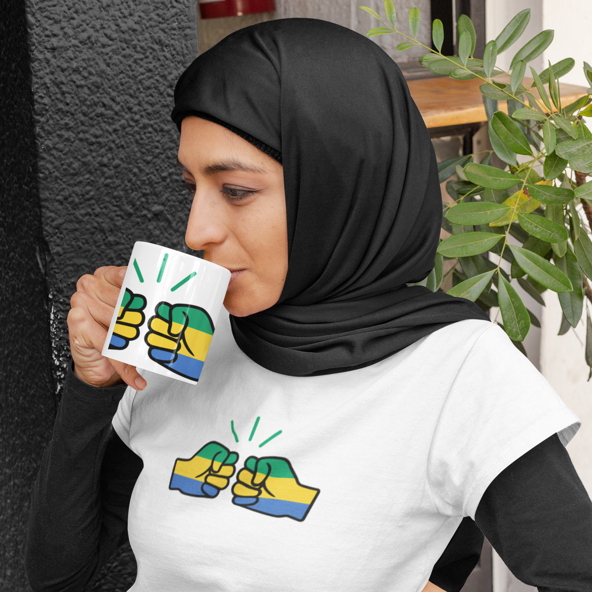 Woman wearing a hijab and white short sleeve t-shirt and holding a coffee mug both displaying the Gabon flag and We Run Tings brand logo