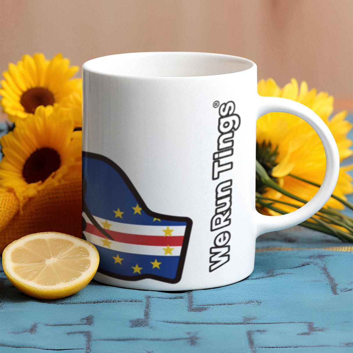 White mug with a We Run Tings logo and Cabo Verde and Cape Verde flag on a wooden surface with yellow flowers and a lemon.