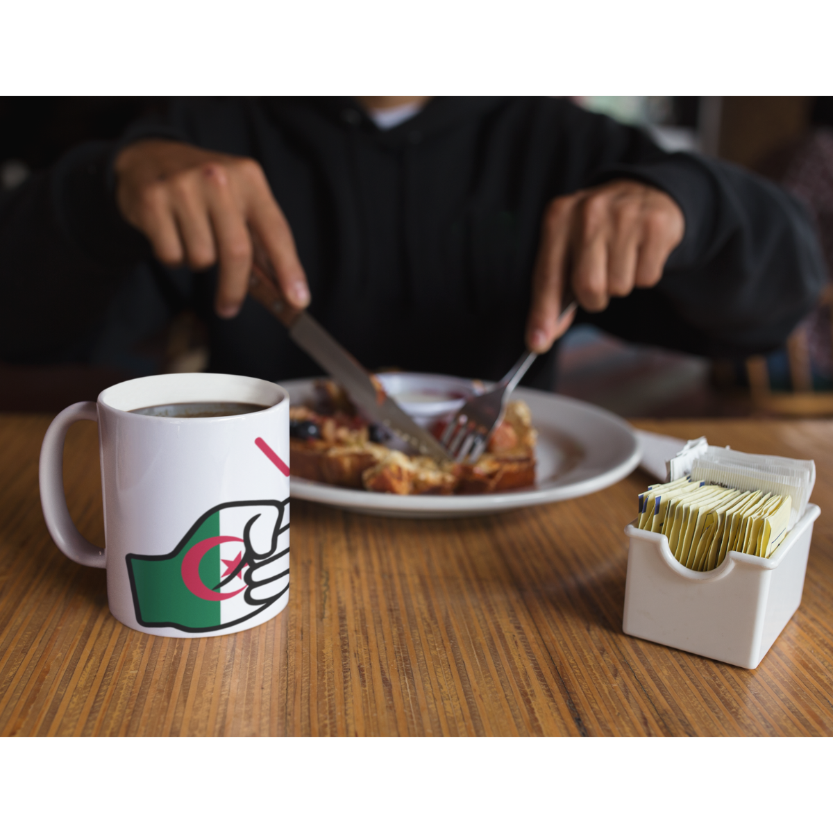 Person eating breakfast with a mug featuring an Algerian flag design on a wooden table.