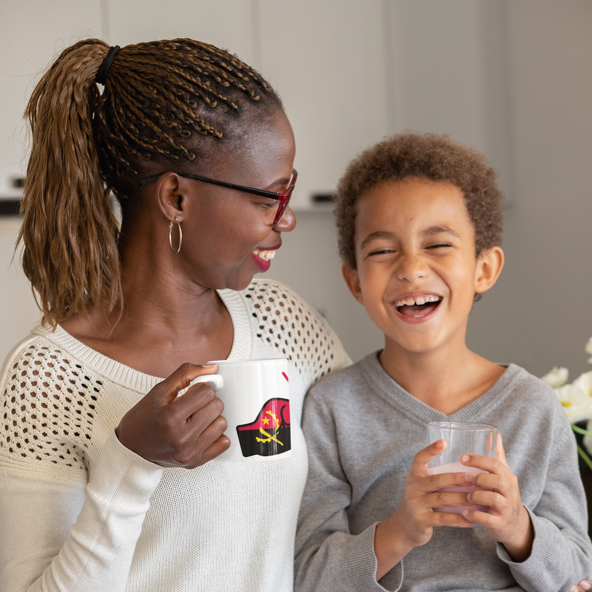 Woman holding a We Run Tings coffee mug displaying the flag of Angola and child sitting together, both holding mugs, with a neutral background
