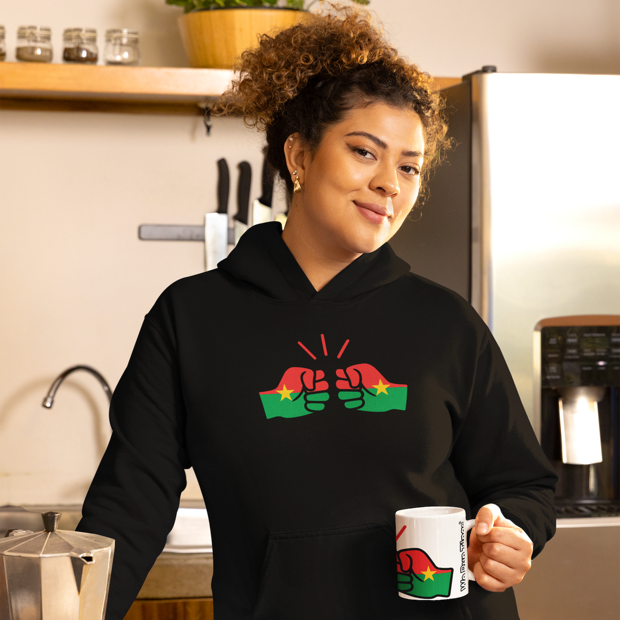 Woman smiling wearing a black We Run Tings hoodie with a colorful design of Burkino Faso, holding a white ceramic mug in a kitchen.