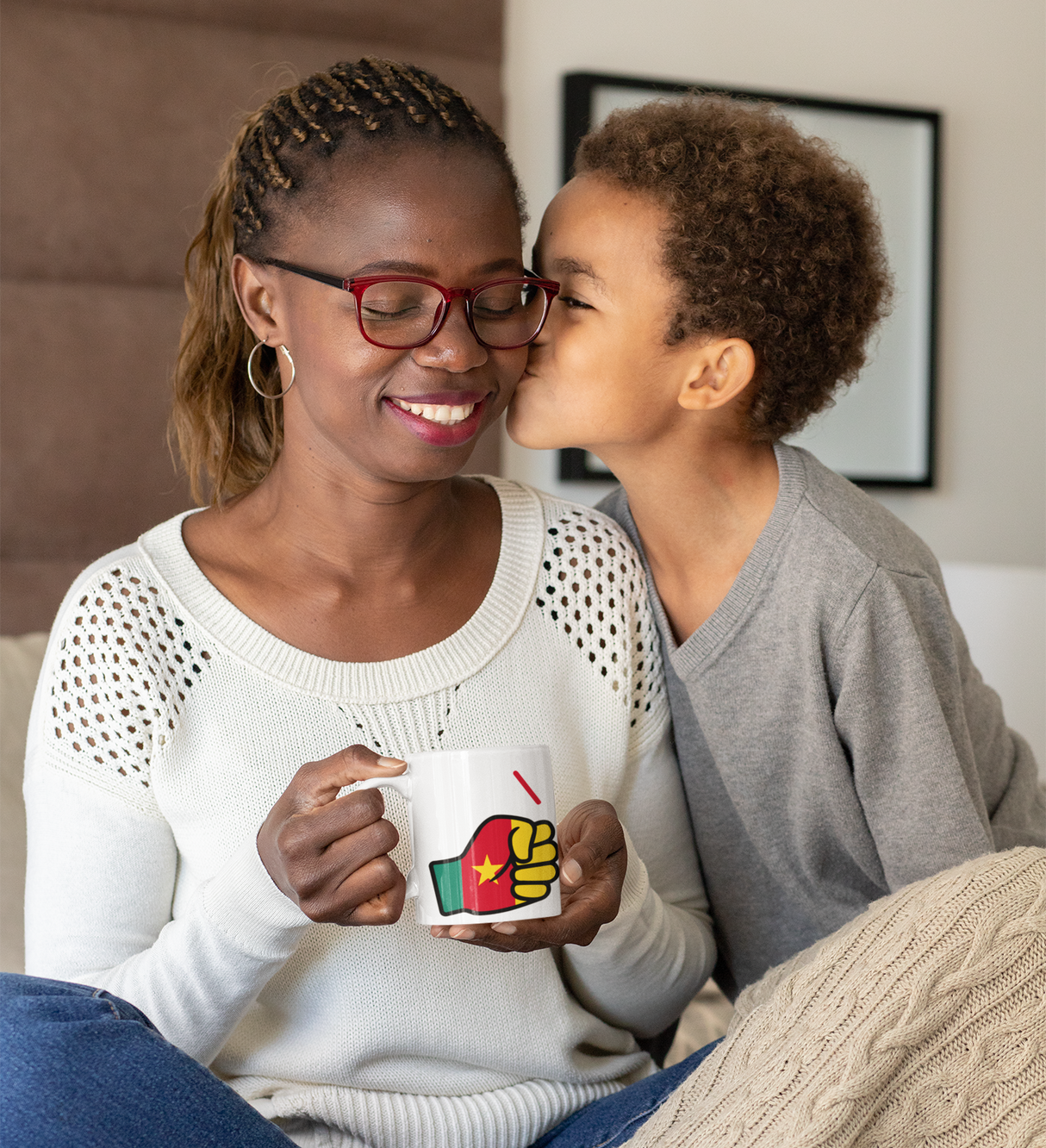 Woman wearing glasses holding a We Run Tings mug, featuring the flag of Cameroon smiling at a child who is kissing her on the cheek.