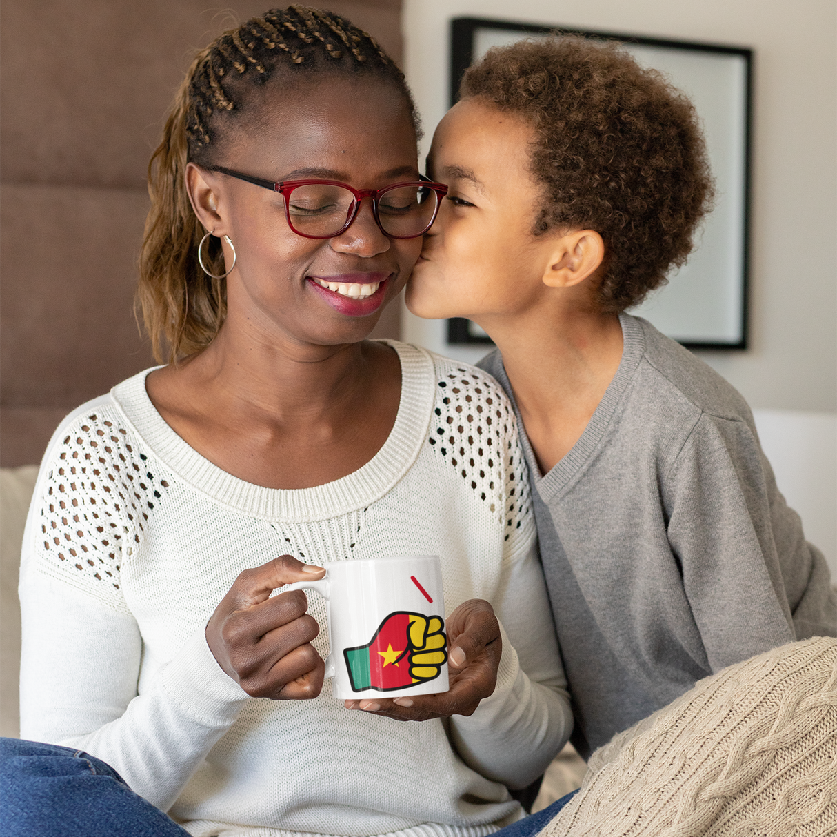 Woman wearing glasses holding a We Run Tings mug, featuring the flag of Cameroon smiling at a child who is kissing her on the cheek.