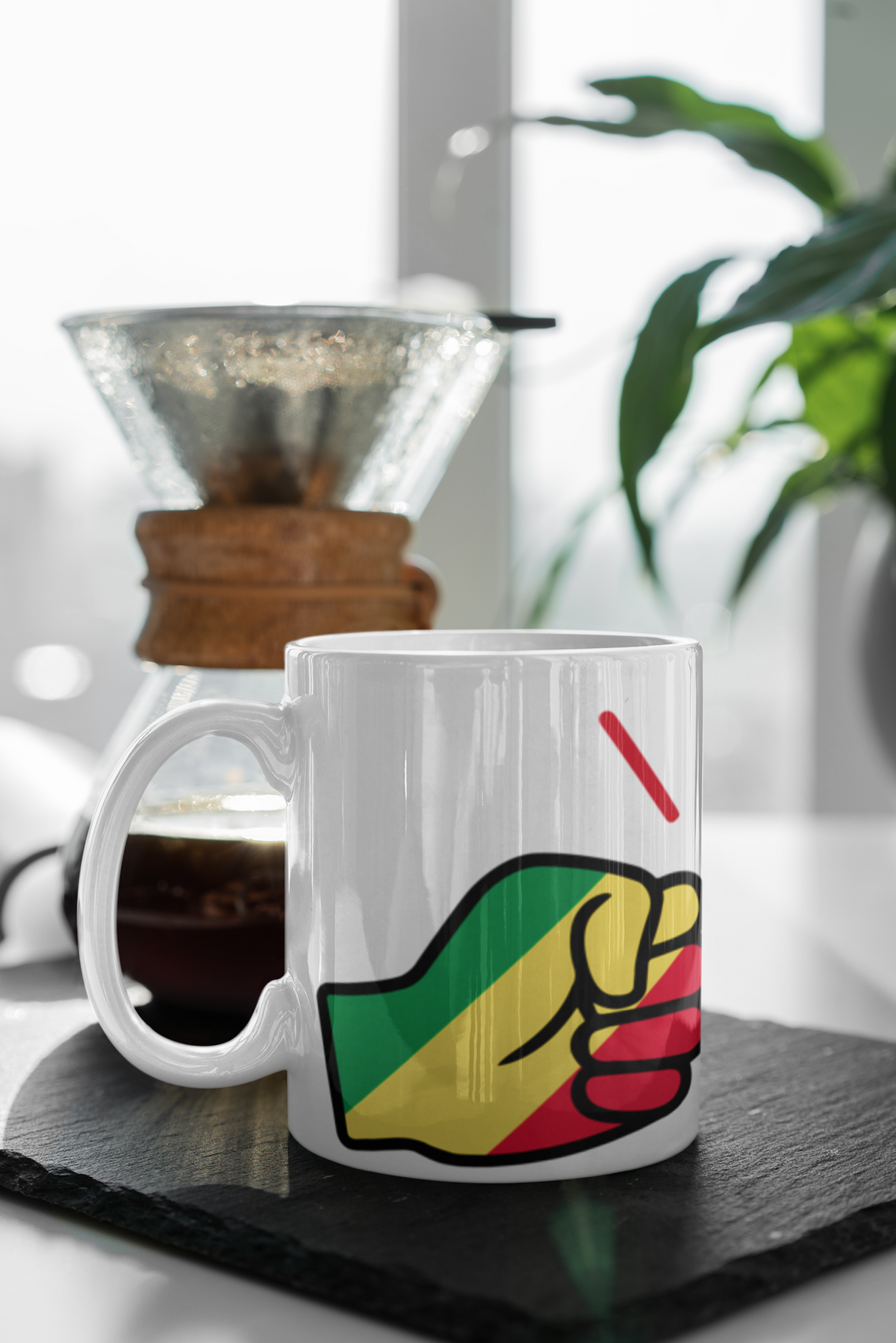 A glossy white ceramic mug on a table next to a coffee maker with a design of We Run Tings Republic of the Congo flag and the brand name. Side view