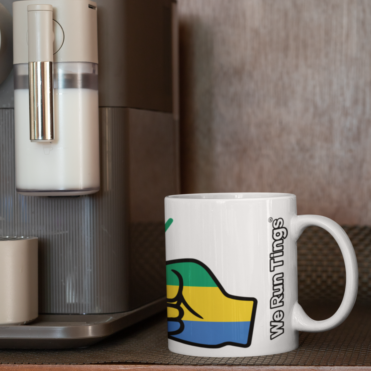 White ceramic We Run Tings Gabon themed mug next to a coffee machine on a wooden surface.