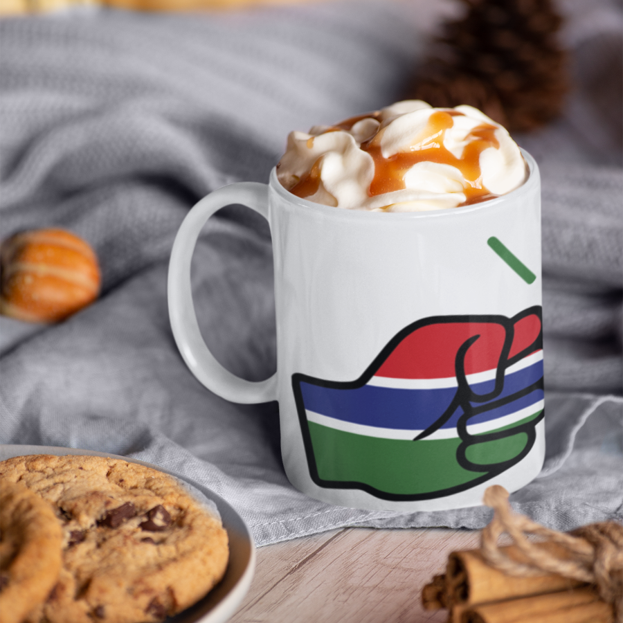 We Run Tings Gambia ceramic mug with colorful design filled with hot chocolate and whipped cream, surrounded by cookies and cinnamon sticks on a wooden surface.