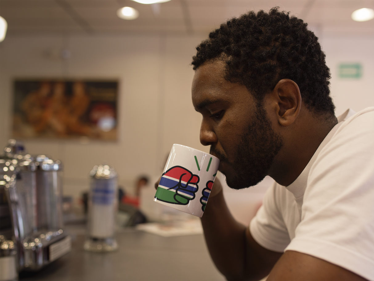 Man drinking from a colorful We Run Tings Gambia themed ceramic mug in a casual setting