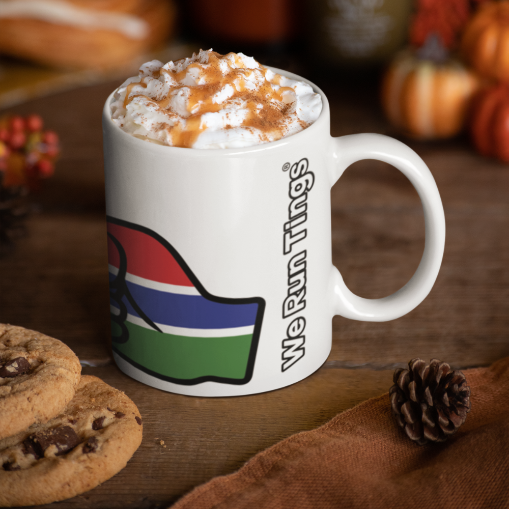 White ceramic We Run Tings Gambia themed mug with colorful design and 'We Run Things' text on a wooden surface with cookies and pumpkins.