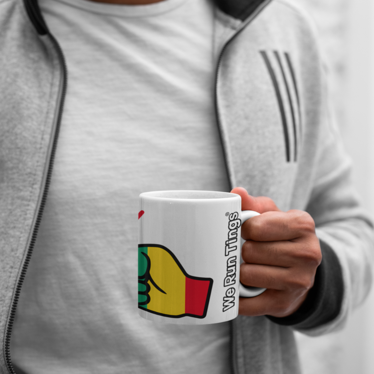 Person holding a We Run Tings Guinea themed mug with a colorful design and text, wearing a gray jacket.