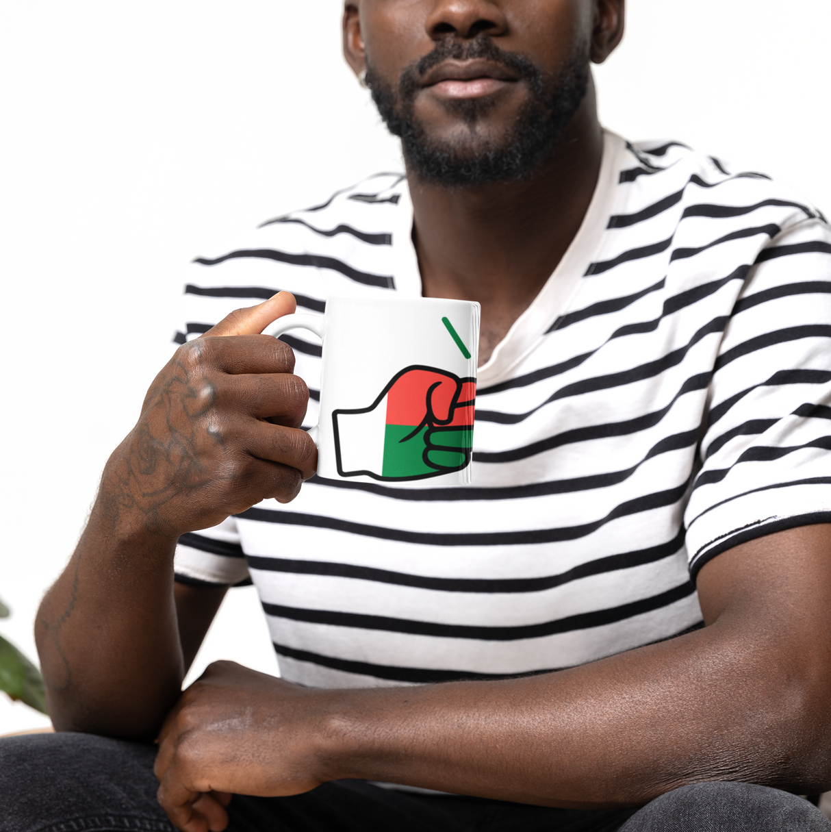Man wearing a striped shirt with a graphic design, sitting on a couch holding a white ceramic We Run Tings Madagascar coffee mug.