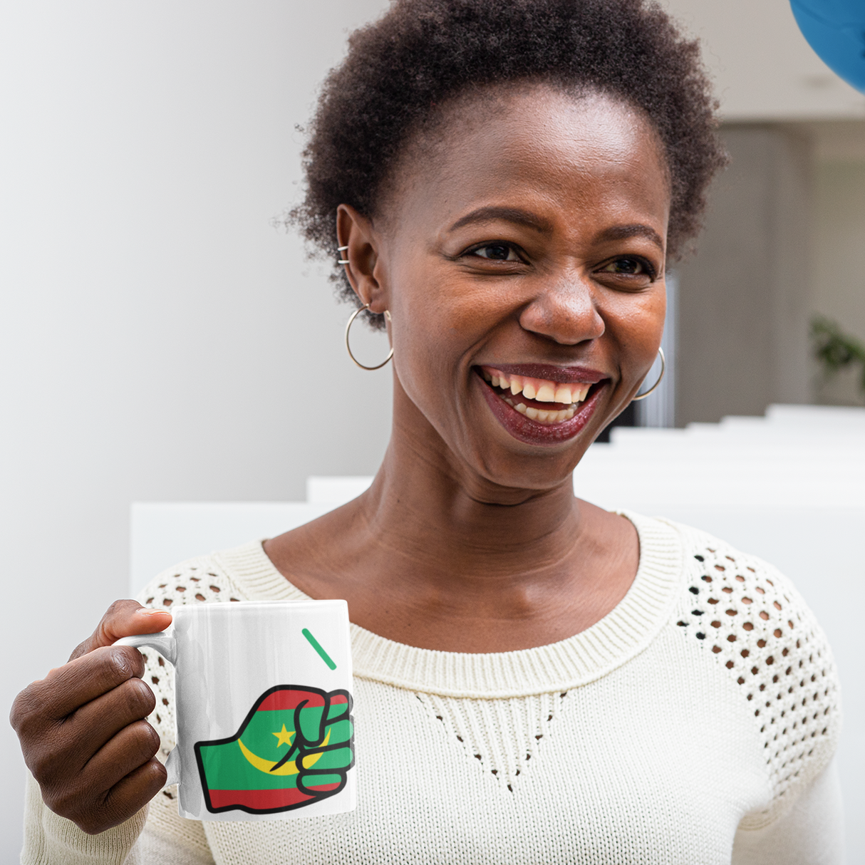 Woman holding a white ceramic We Run Tings Mauritania mug, cup with a green and red fist design, smiling indoors.