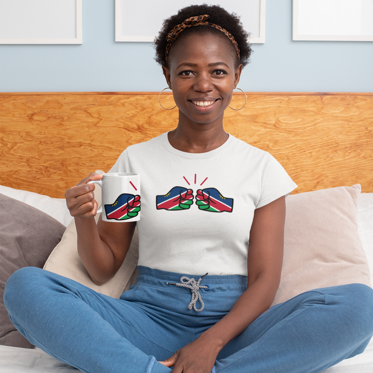 Woman sitting on a bed holding a We Run Tings Namibia themed mug with a matching design, wearing a white t-shirt with a colorful We Run Tings graphic logo