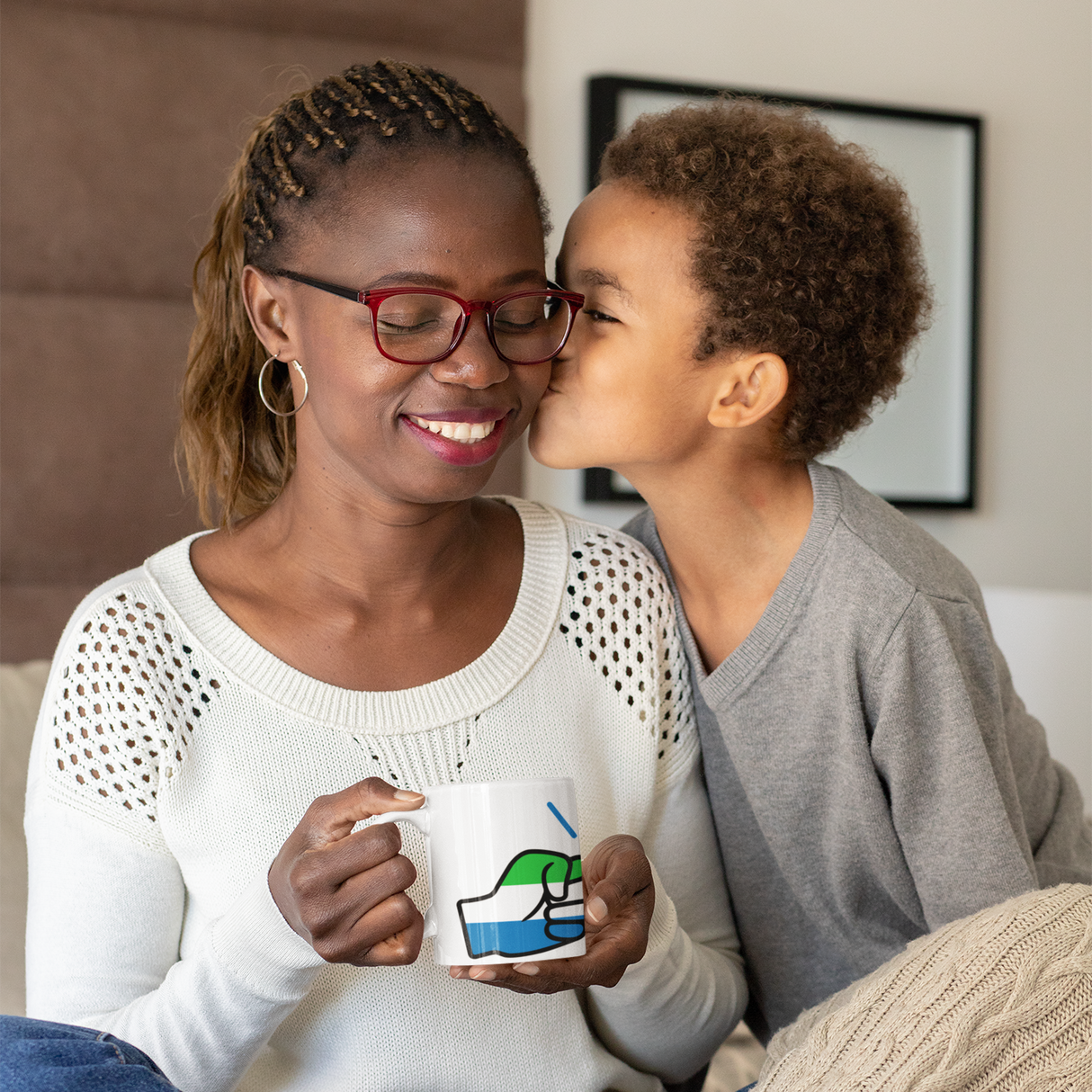 Woman with glasses holding a Sierra Leone We Run Tings white ceramic coffee, tea mug, smiling at a child who is kissing her on the cheek.