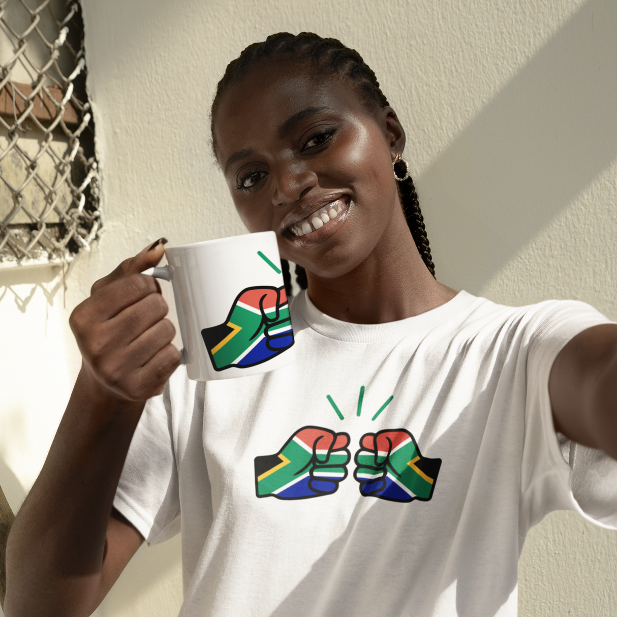 A smiling Woman wearing a white t-shirt with colorful South Africa We Run Tings graphic design, and white ceramic coffee mug standing indoors.