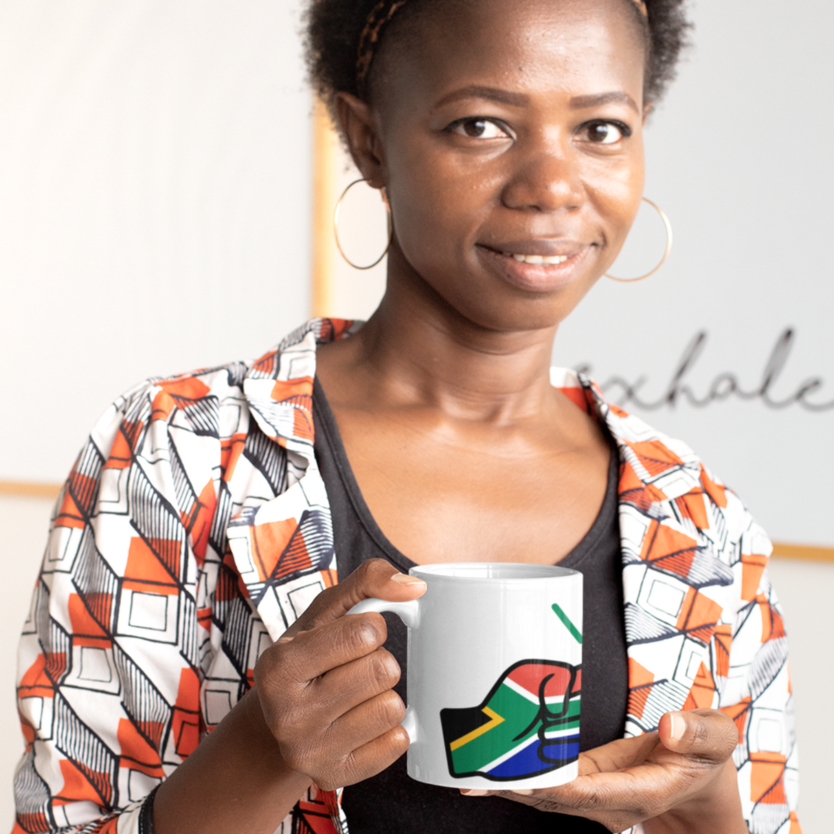 Woman smiling, holding a white ceramic We Run Tings, South Africa themed mug with the flag within the design, wearing a patterned jacket.