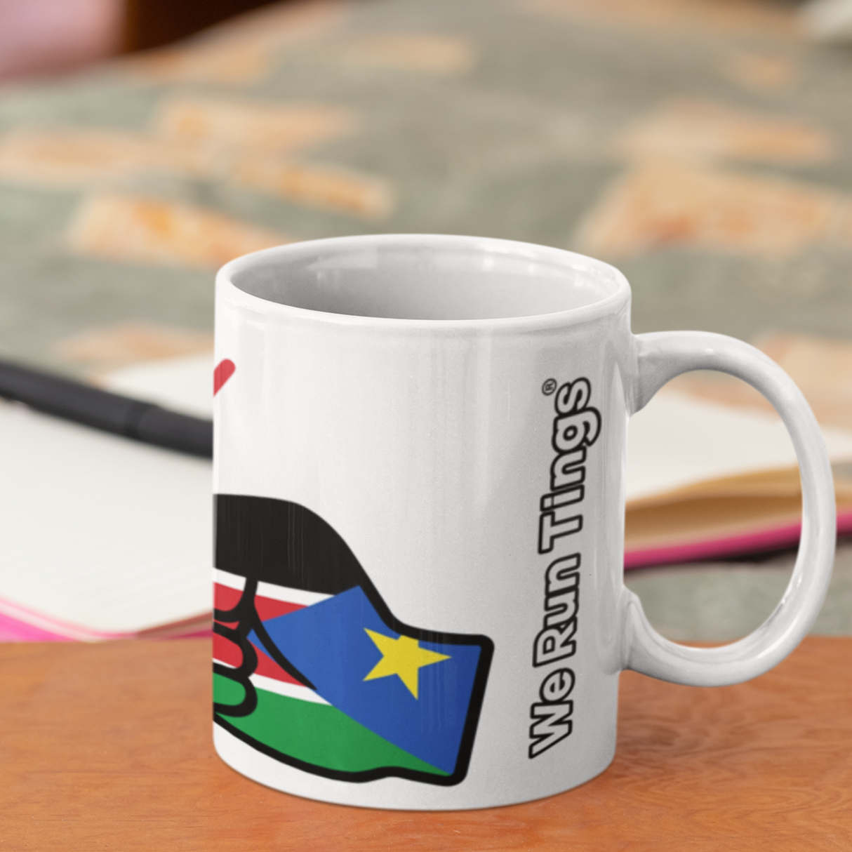 We Run Tings White mug with a colorful design featuring the flag of South Sudan and text on a wooden surface
