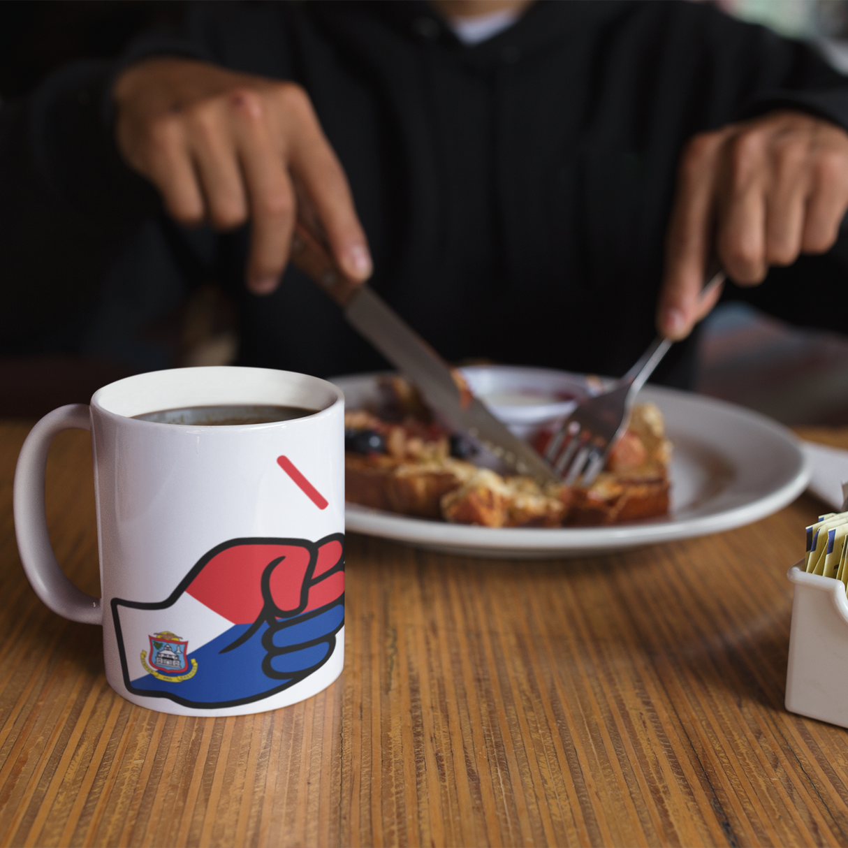 Person eating breakfast with a We Run Tings, St. Martin Mug featuring a colorful design on a wooden table.