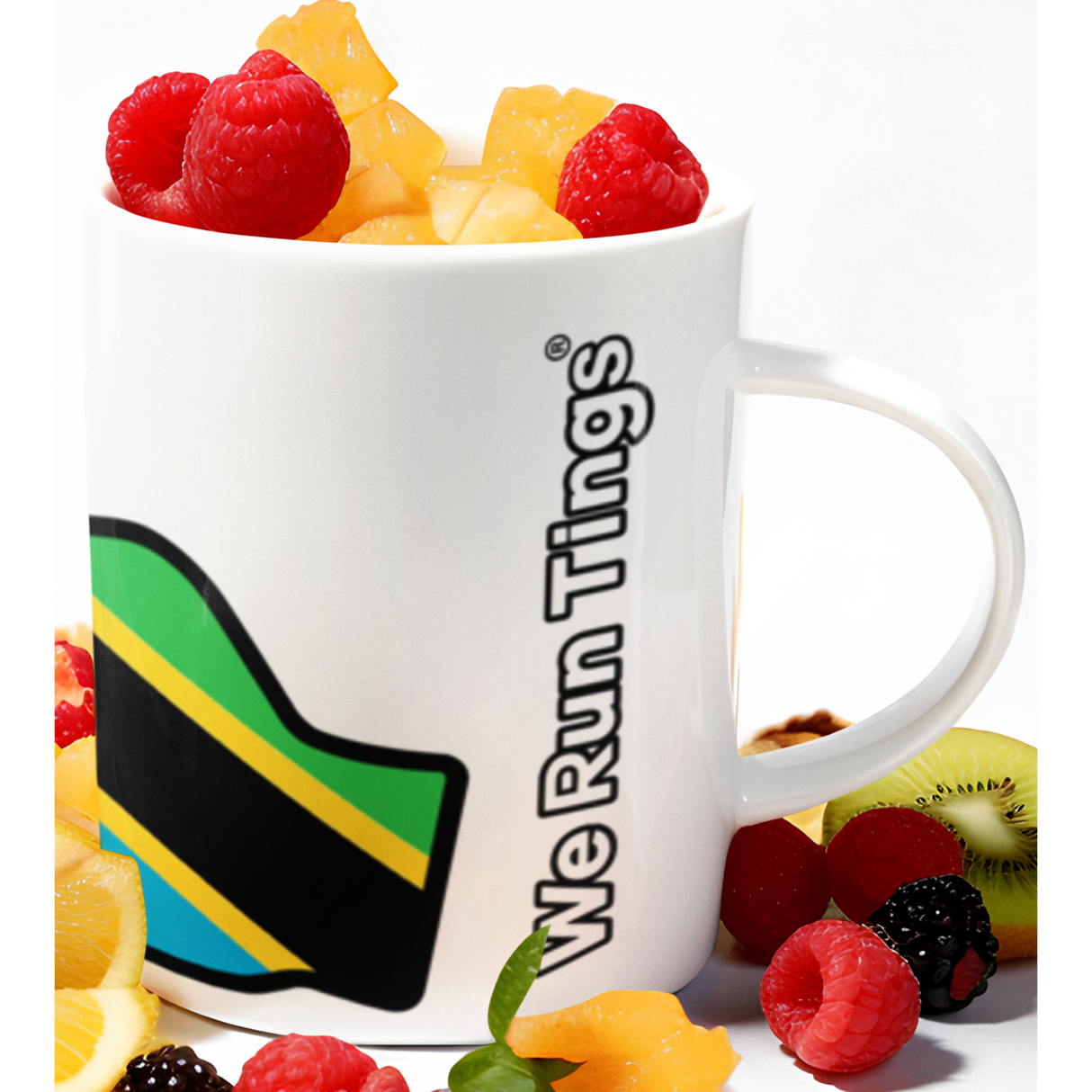 White ceramic mug featuring the “We Run Tings” logo with the Tanzania flag design, surrounded by fresh fruit including raspberries, blackberries, orange slices, kiwi, and diced mango, styled on a clean white background.