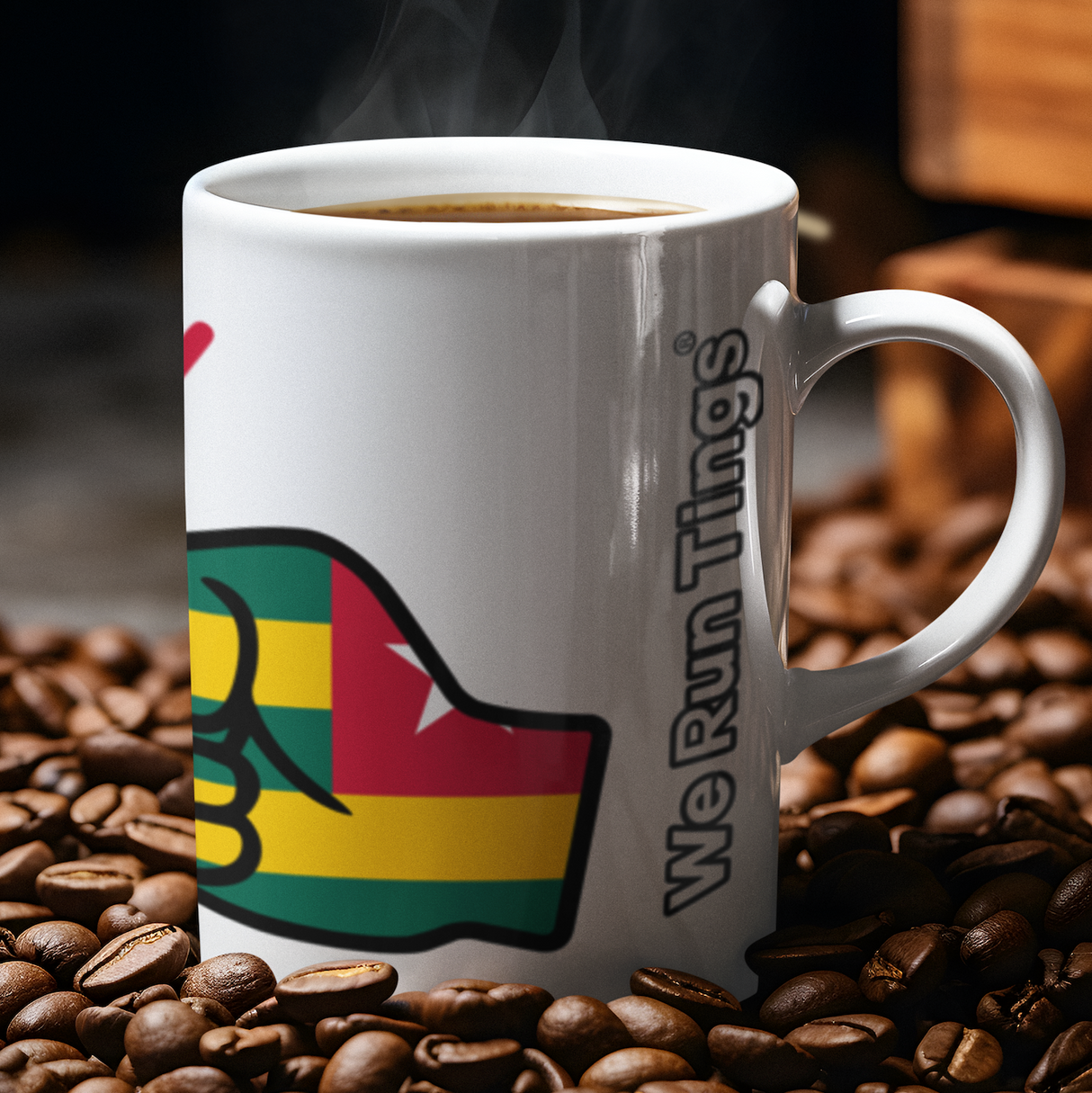 White ceramic “We Run Tings” Togo mug filled with hot coffee, surrounded by roasted coffee beans, featuring a raised fist design in Togo flag colors with visible steam rising in a warm, rich setting.