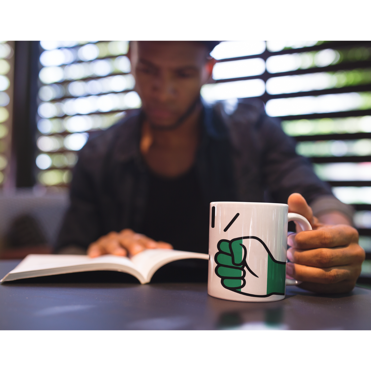 Man holding a We Run Tings Nigeria white, ceramic, mug, cup with a fist symbol while reading a book in a blurred indoor setting
