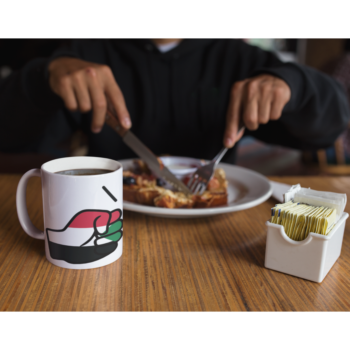 Person eating breakfast with a White We Run Tings, Sudan Mug with a graphic of a fist on a wooden table.