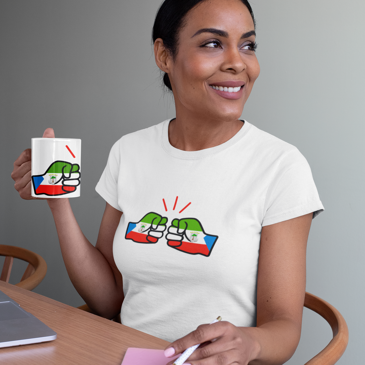 Woman wearing a white t-shirt with a We Run Tings Equatorial Guinea logo and holding a matching ceramic mug, and sitting at a table.