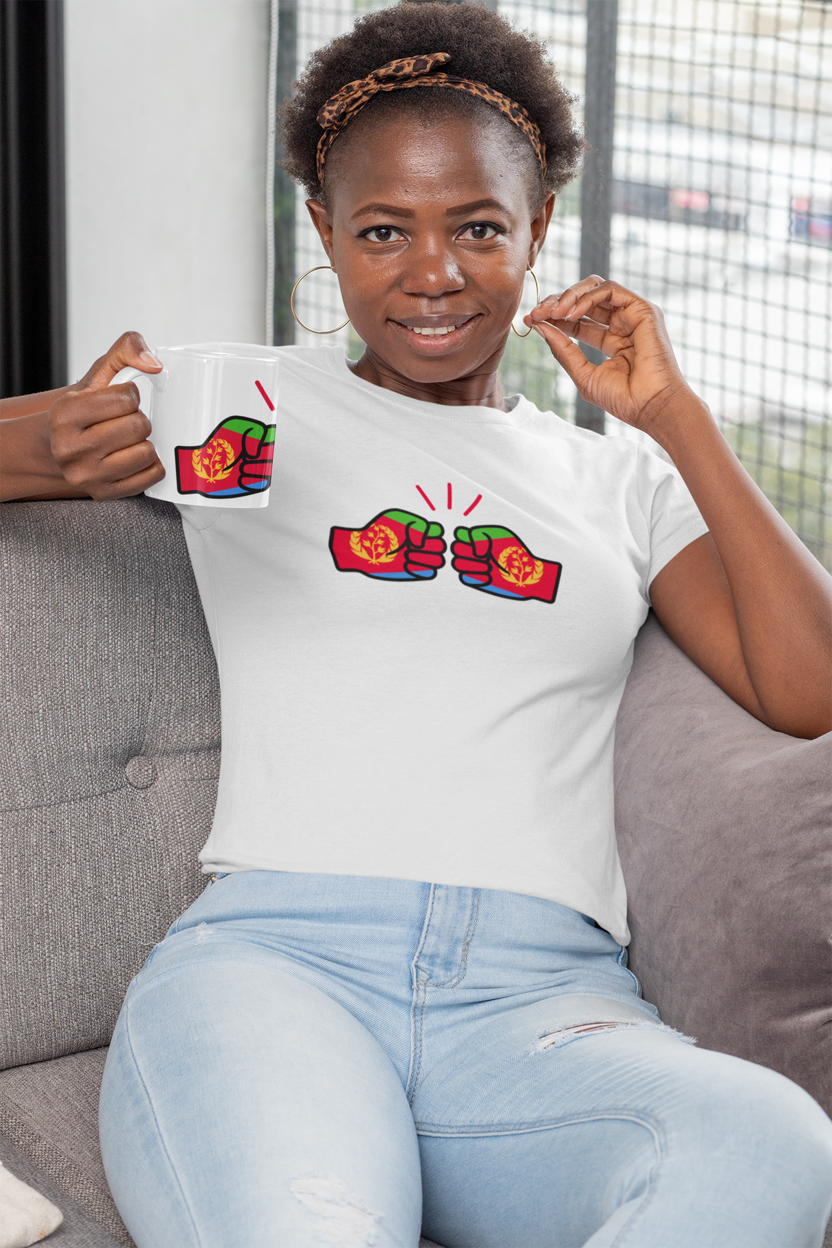 Woman wearing a white We Run Tings Eritrea Mug and t-shirt with colorful graphics, sitting on a couch.