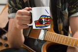 Person holding a Glossy white ceramic mug featuring a patriotic We Run Tings logo with two fists displaying the flag of Eswatini in front of an acoustic guitar
