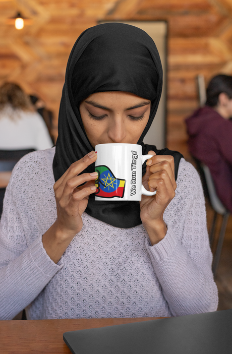 Woman wearing a hijab drinking from a We Run Tings white ceramic Ethiopia mug with a colorful design, sitting in a casual setting.