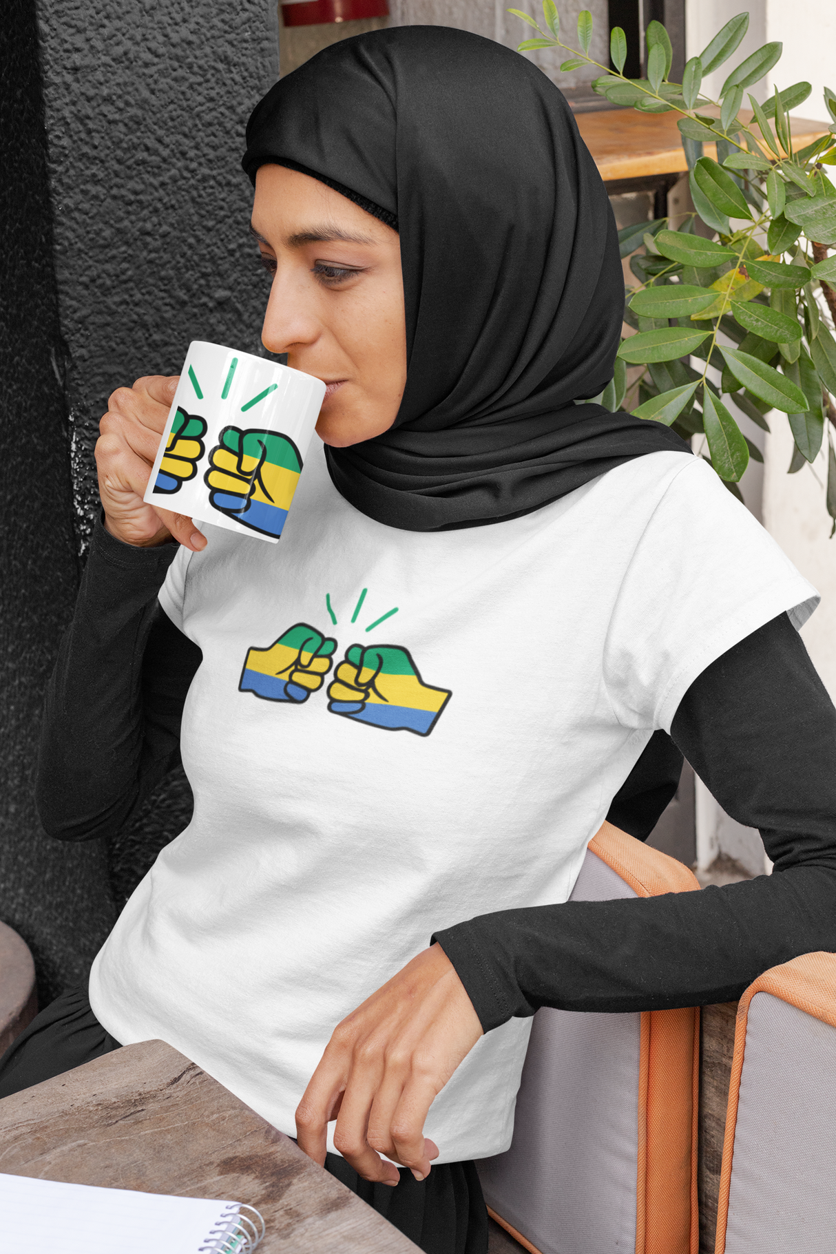 Woman wearing a hijab and white short sleeve t-shirt and holding a coffee mug both displaying the Gabon flag and We Run Tings brand logo