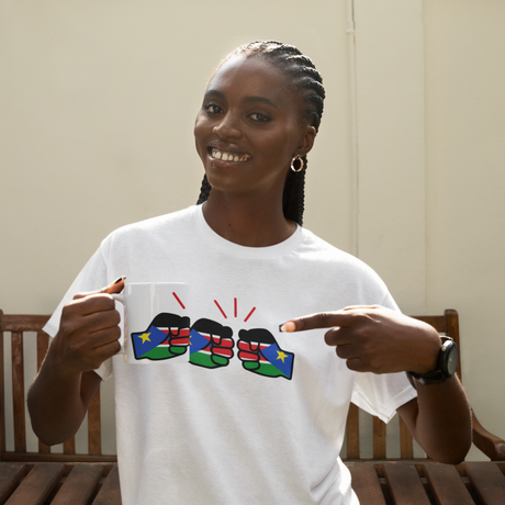 Woman smiling pointing to a white t-shirt with colorful graphic design of We Run Tings themed logo of South Sudan and matching white ceramic coffee, tea Mug