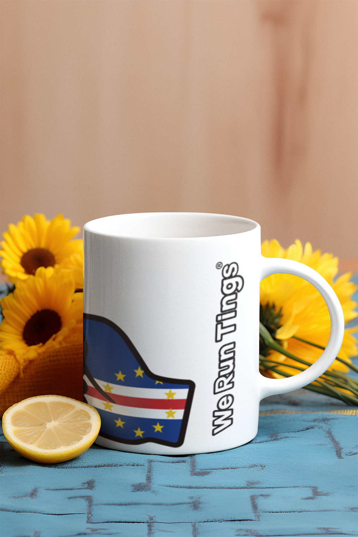 White mug with a We Run Tings logo and Cabo Verde and Cape Verde flag on a wooden surface with yellow flowers and a lemon.