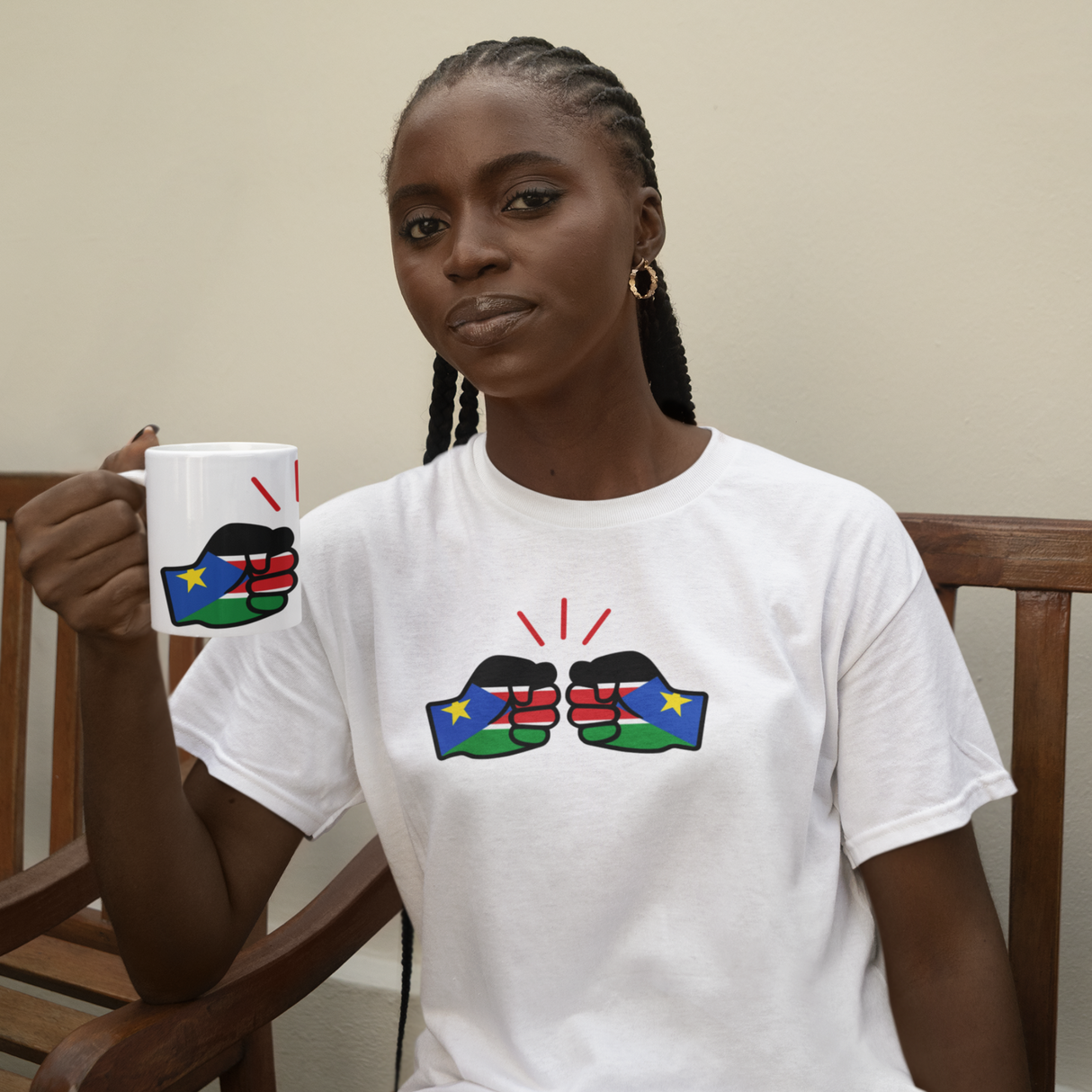 Woman sitting on a bench holding a white ceramic We Run Tings South Sudan themed mug, wearing a matching white t-shirt with colorful designs.