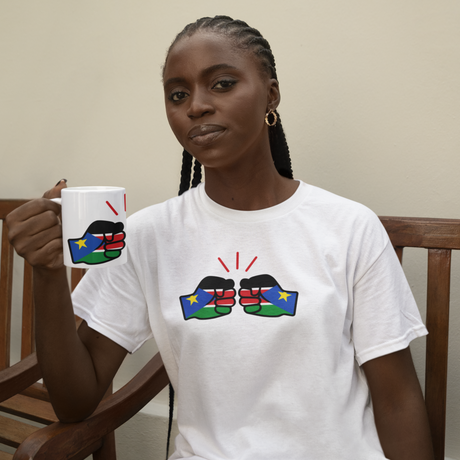 Woman sitting on a bench holding a white ceramic We Run Tings South Sudan themed mug, wearing a matching white t-shirt with colorful designs.