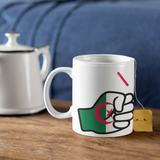 White mug with a design of the flag of Algeria on a wooden surface next to a teapot and tea bag.