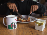 Person eating breakfast with a mug featuring an Algerian flag design on a wooden table.