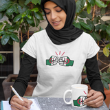 Woman wearing a white We Run Tings Algerian themed t-shirt with a graphic design, sitting at a table with a white ceramic coffee mug cup displaying the same logo