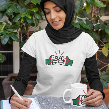 Woman wearing a white We Run Tings Algerian themed t-shirt with a graphic design, sitting at a table with a white ceramic coffee mug cup displaying the same logo