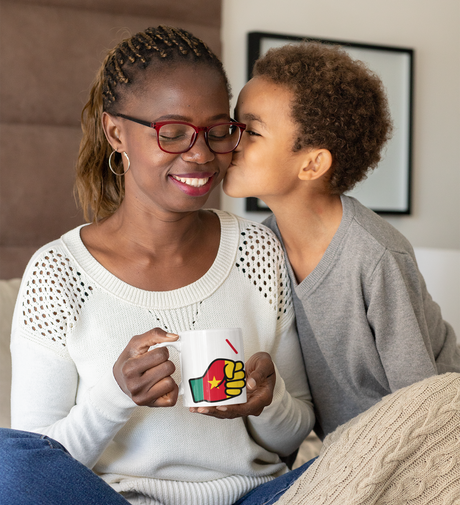 Woman wearing glasses holding a We Run Tings mug, featuring the flag of Cameroon smiling at a child who is kissing her on the cheek.