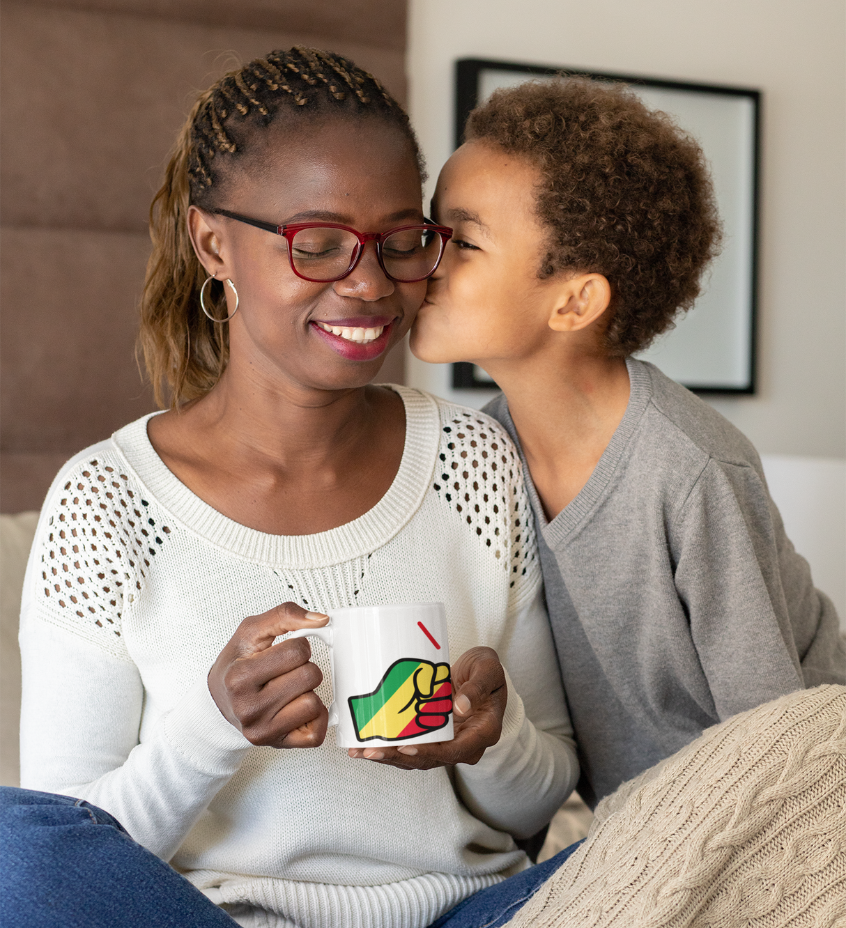 Woman with glasses holding a We Run Tings, Republic of the Congo themed mug and a child kissing her on the cheek, indoor setting