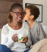 Woman with glasses holding a We Run Tings, Republic of the Congo themed mug and a child kissing her on the cheek, indoor setting