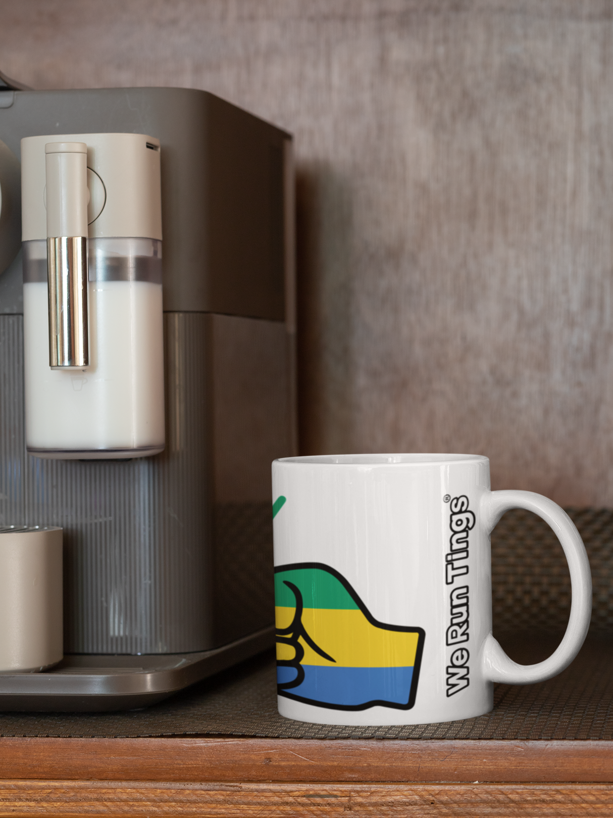 White ceramic We Run Tings Gabon themed mug next to a coffee machine on a wooden surface.