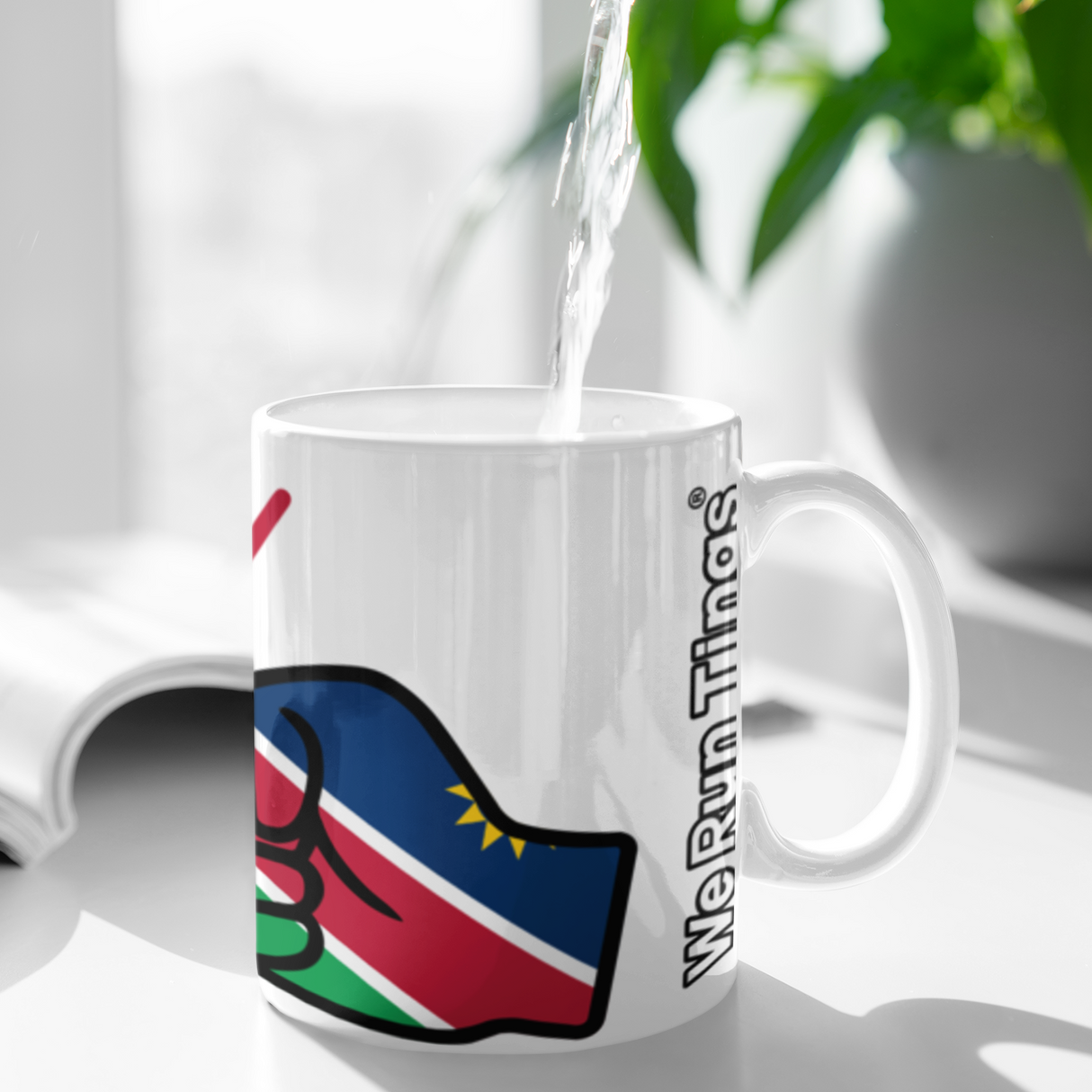 White mug with a We Run Tings Namibia logo being filled with water, placed on a white surface with a plant in the background.