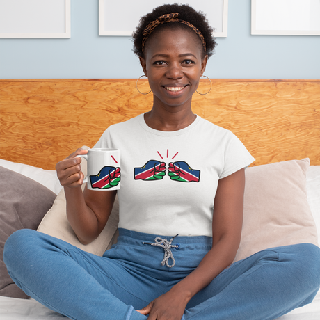 Woman sitting on a bed holding a We Run Tings Namibia themed mug with a matching design, wearing a white t-shirt with a colorful We Run Tings graphic logo