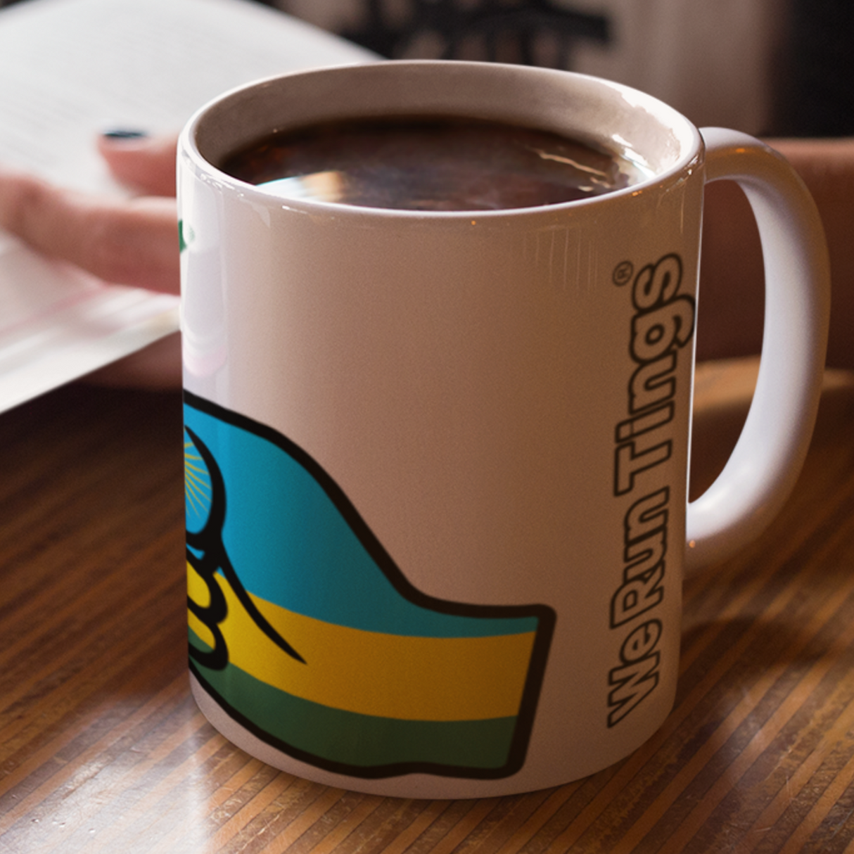 We Run Tings white ceramic tea, coffee cup, mug with a colouful Rwanda flag design and text on a wooden surface, next to an open book. Cup full of coffee.