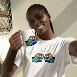 A smiling Woman wearing a white t-shirt with colorful South Africa We Run Tings graphic design, and white ceramic coffee mug standing indoors.