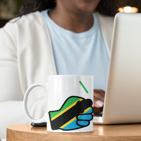 Lady using a laptop sitting at a table featuring a white We Run Tings, Tanzania tea, coffee ceramic mug, cup featuring the flag of Tanzania within the We Run Tings fist logo