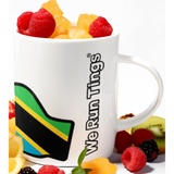 White ceramic mug featuring the “We Run Tings” logo with the Tanzania flag design, surrounded by fresh fruit including raspberries, blackberries, orange slices, kiwi, and diced mango, styled on a clean white background.